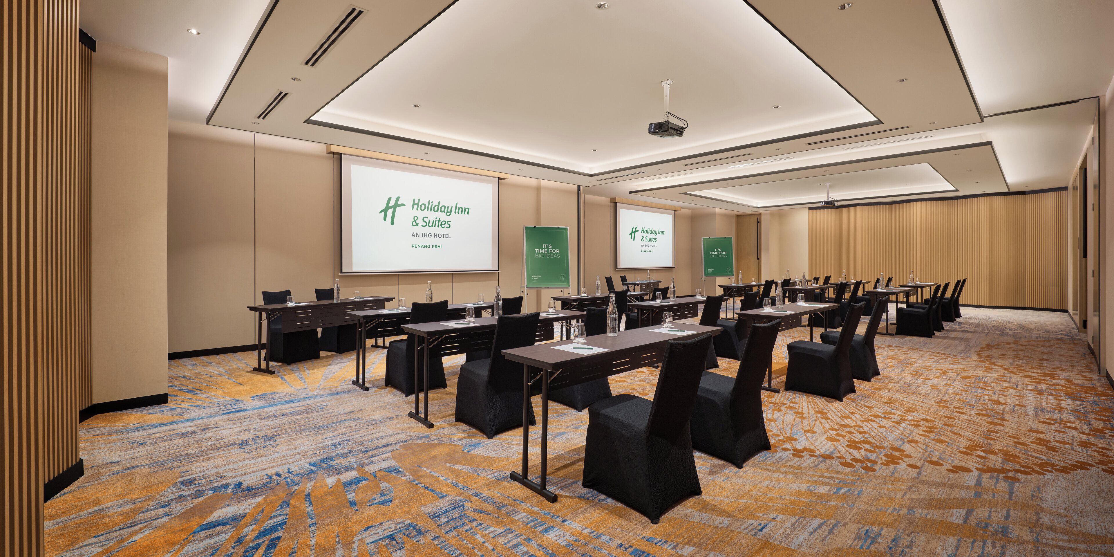 Une salle de conférence dotée de plusieurs tables et chaises, d’un écran de projection affichant le logo « Holiday Inn & Suites », et d’un sol recouvert de moquette.