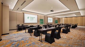 Une salle de conférence dotée de plusieurs tables et chaises, d’un écran de projection affichant le logo « Holiday Inn & Suites », et d’un sol recouvert de moquette.