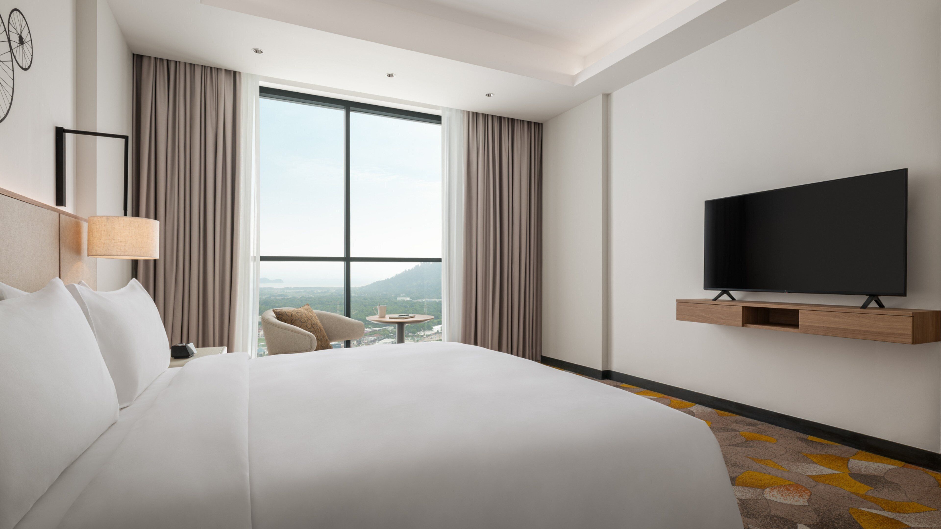 Quarto de hotel com uma cama grande, uma TV em uma prateleira de madeira, uma cadeira, uma mesinha e vista da cidade através de grandes janelas.