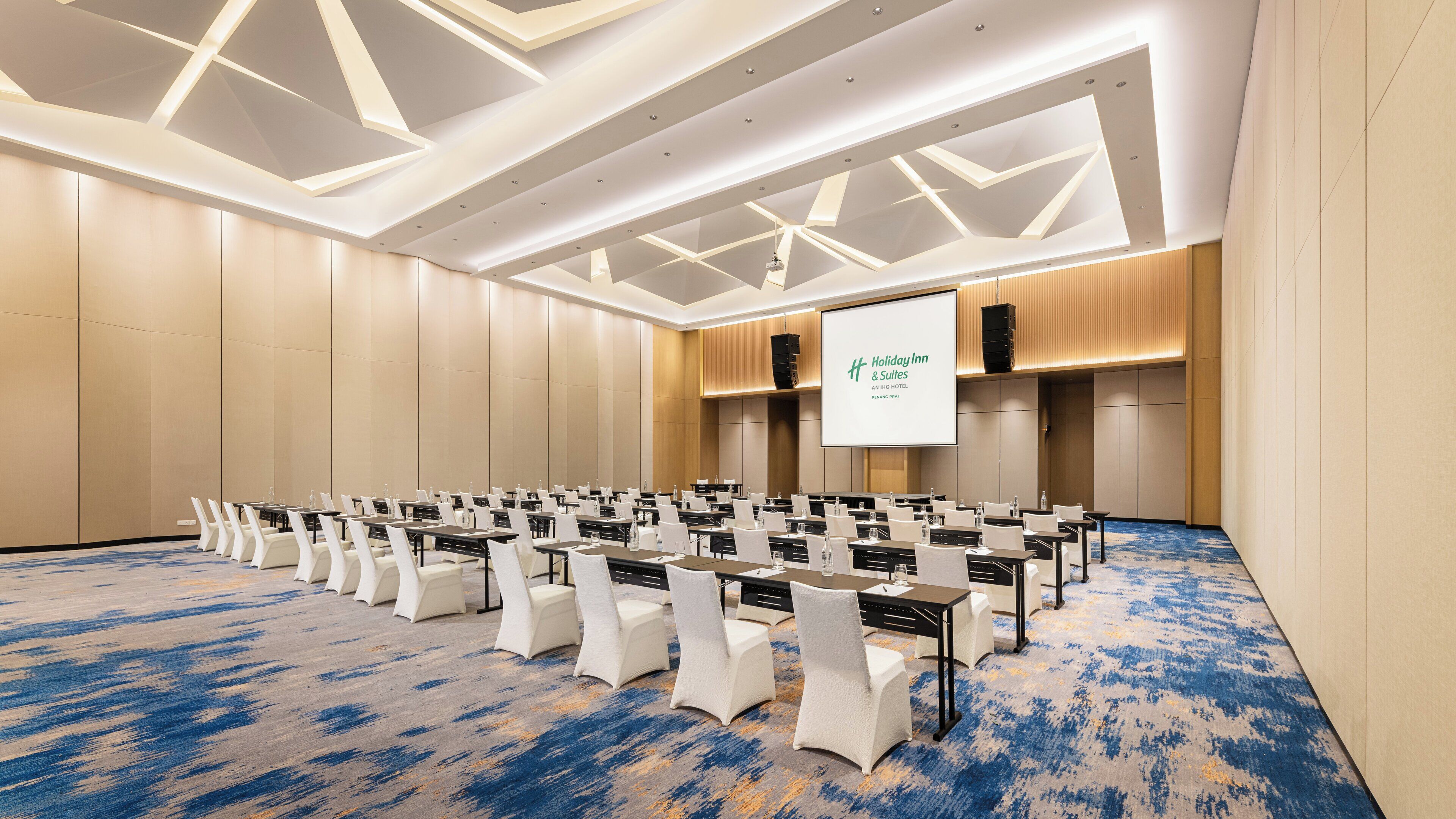 A conference room with rows of tables and chairs, a large screen, and a modern ceiling design.