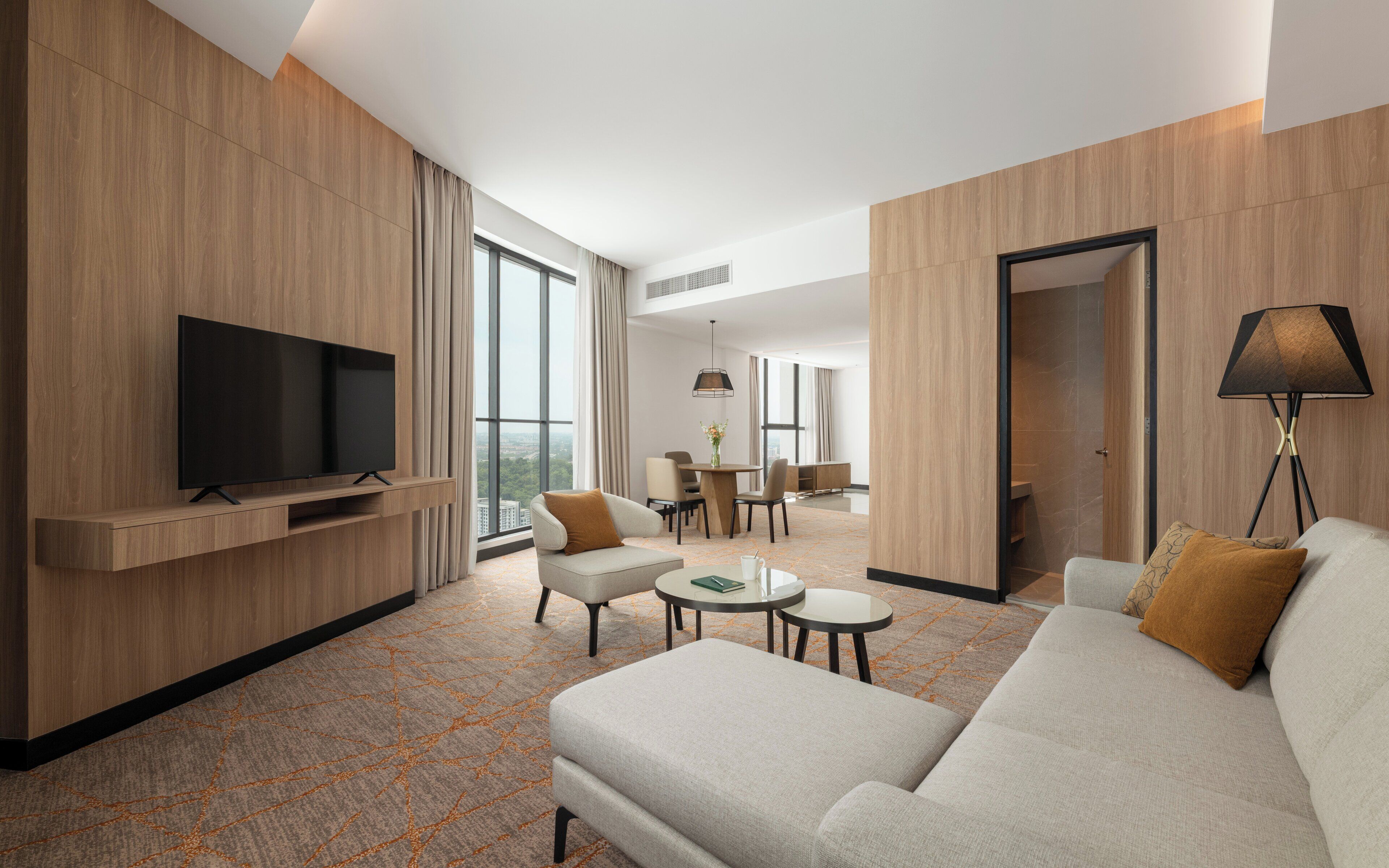 A modern hotel room with a flat-screen TV, a sofa, a dining area, and a large window offering a city view.