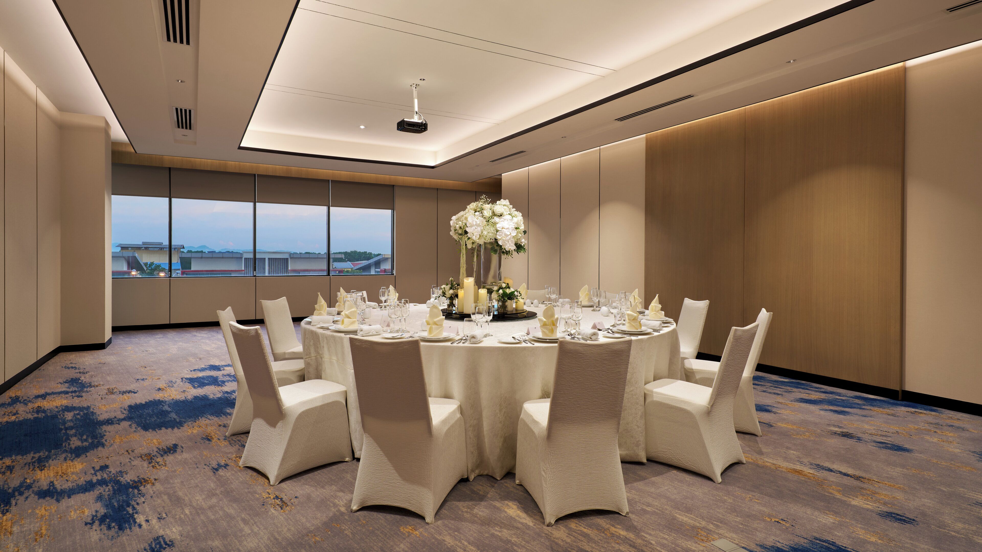 A conference room with a round table set for a formal event, large windows with a view, and a carpeted floor.