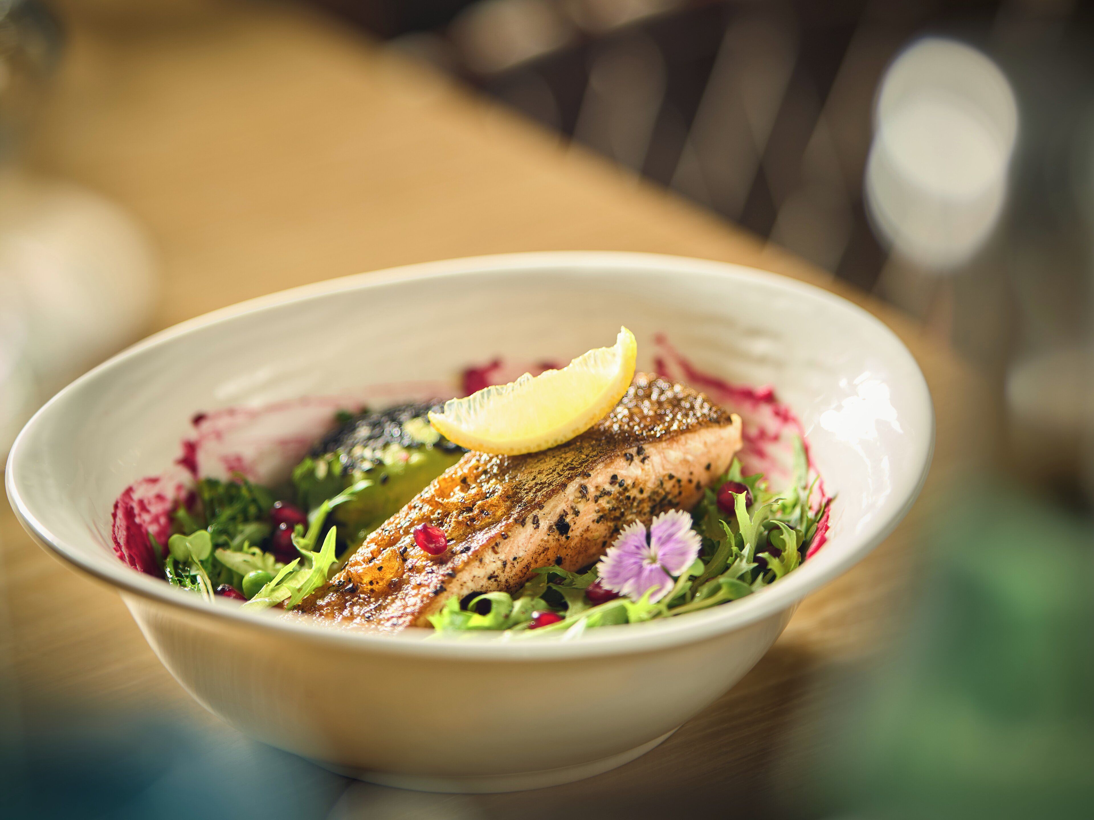 A bowl of food with a piece of fish, greens, and a lemon slice.