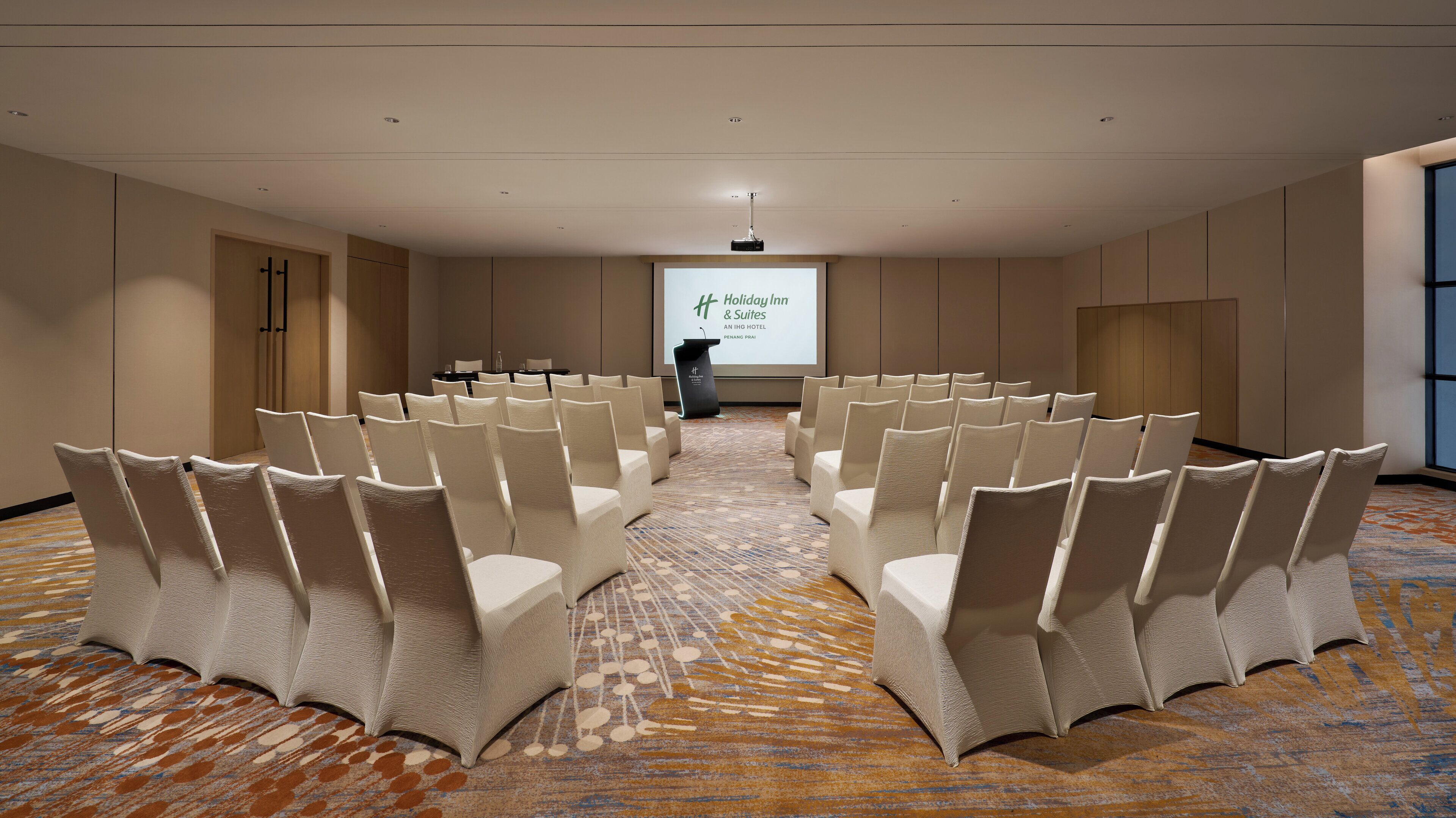 Un salón de conferencias con filas de sillas blancas, un atril y una pantalla proyectora que muestra el logotipo de Holiday Inn & Suites.