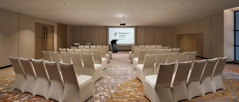 Un salón de conferencias con filas de sillas blancas, un atril y una pantalla proyectora que muestra el logotipo de Holiday Inn & Suites.