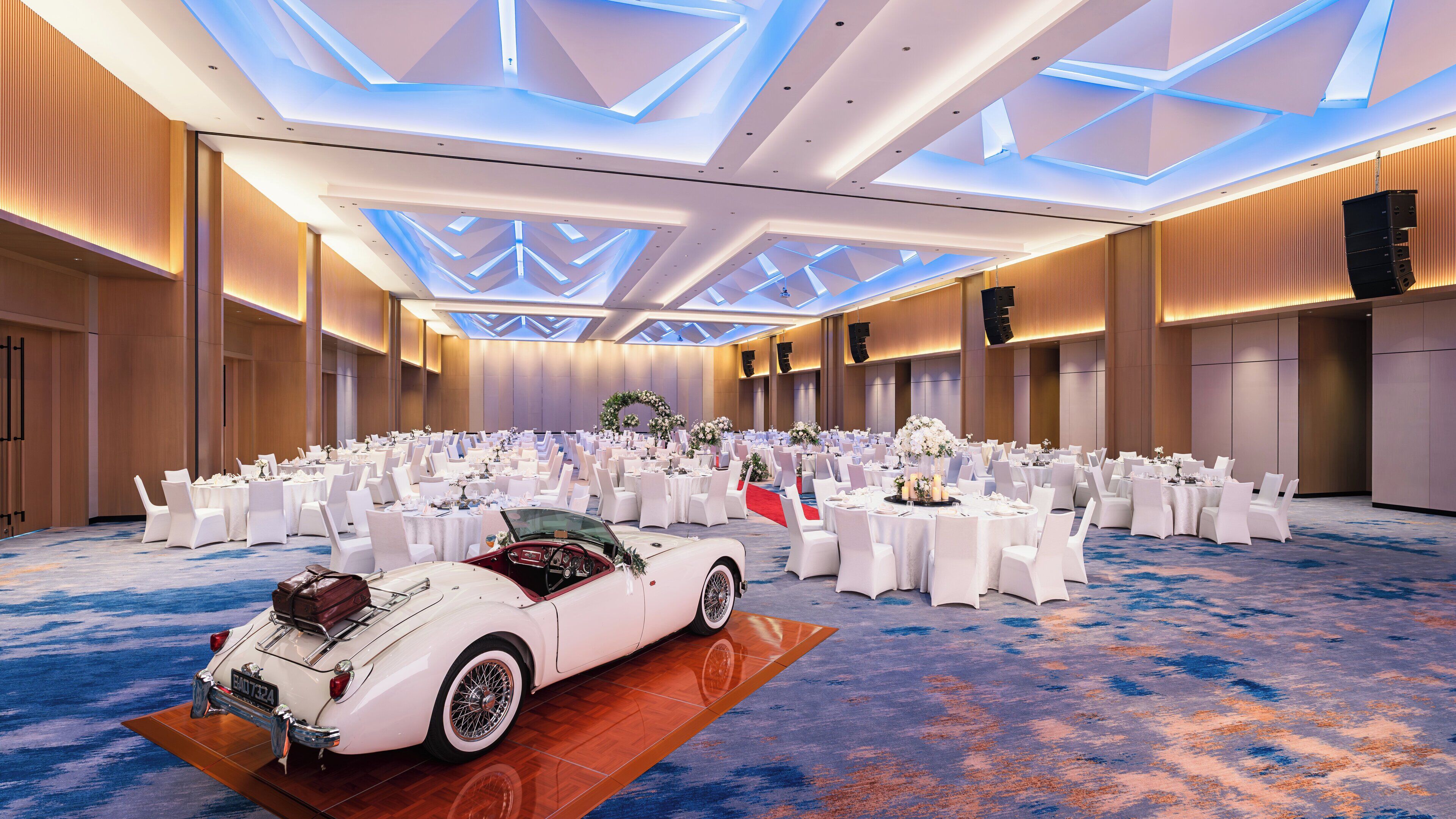 A spacious banquet hall with round tables, white chairs, and a classic car display.