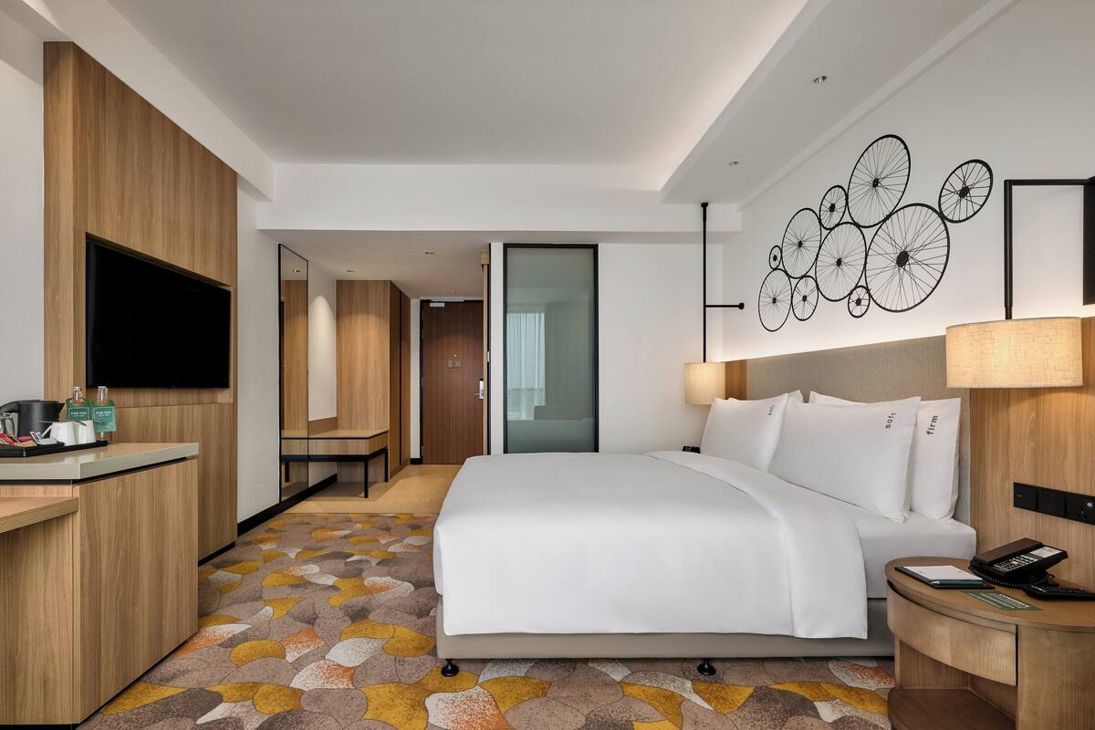 Une chambre d’hôtel moderne dotée d’un grand lit, d’un bureau avec un téléviseur à écran plat, d’une table de chevet avec une lampe et d’une œuvre d’art représentant des bicyclettes, accrochée au mur.