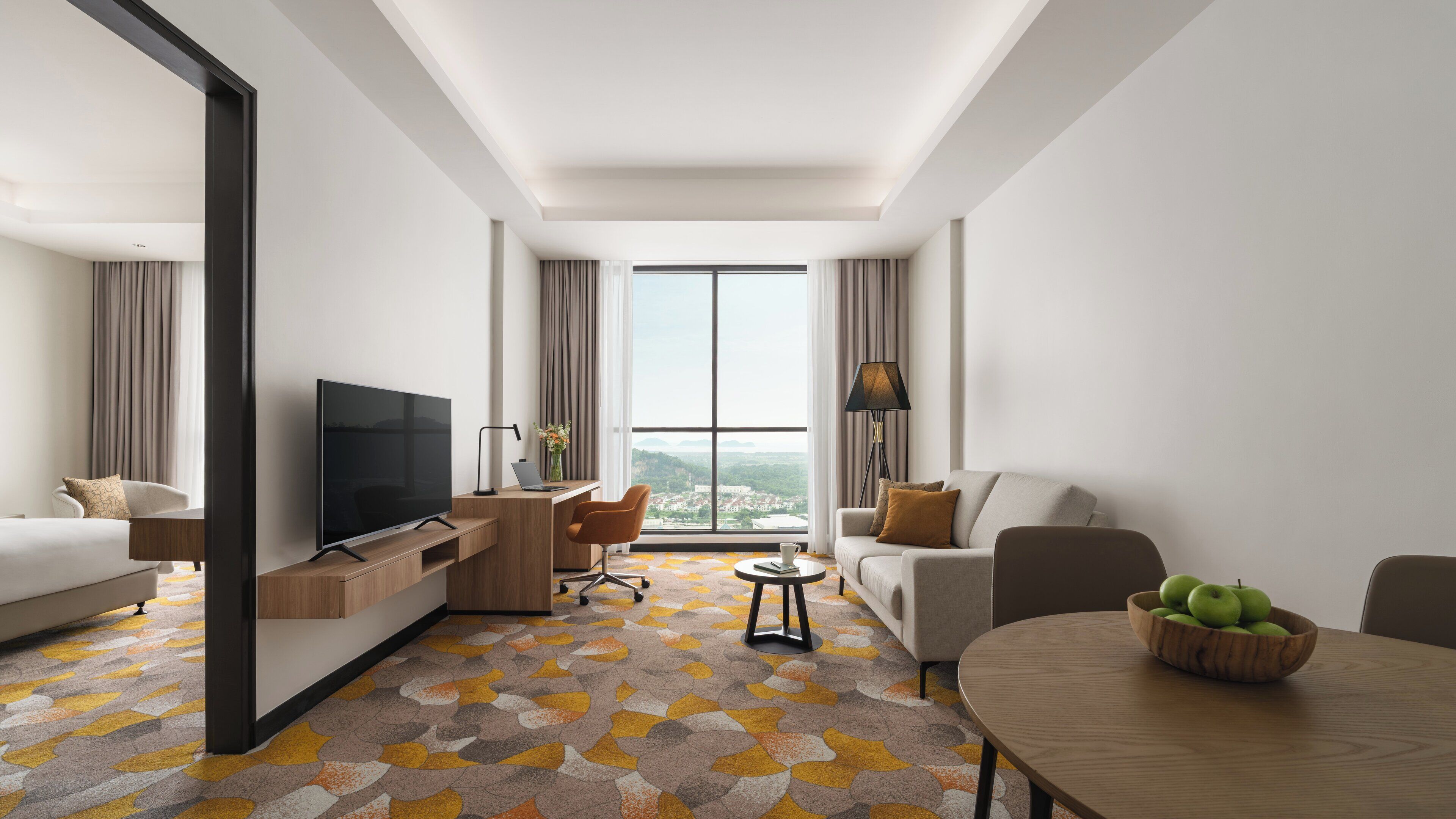 A modern hotel room with a large TV, a desk with a laptop, a sofa, a dining table with apples, and a view of the city through large windows.