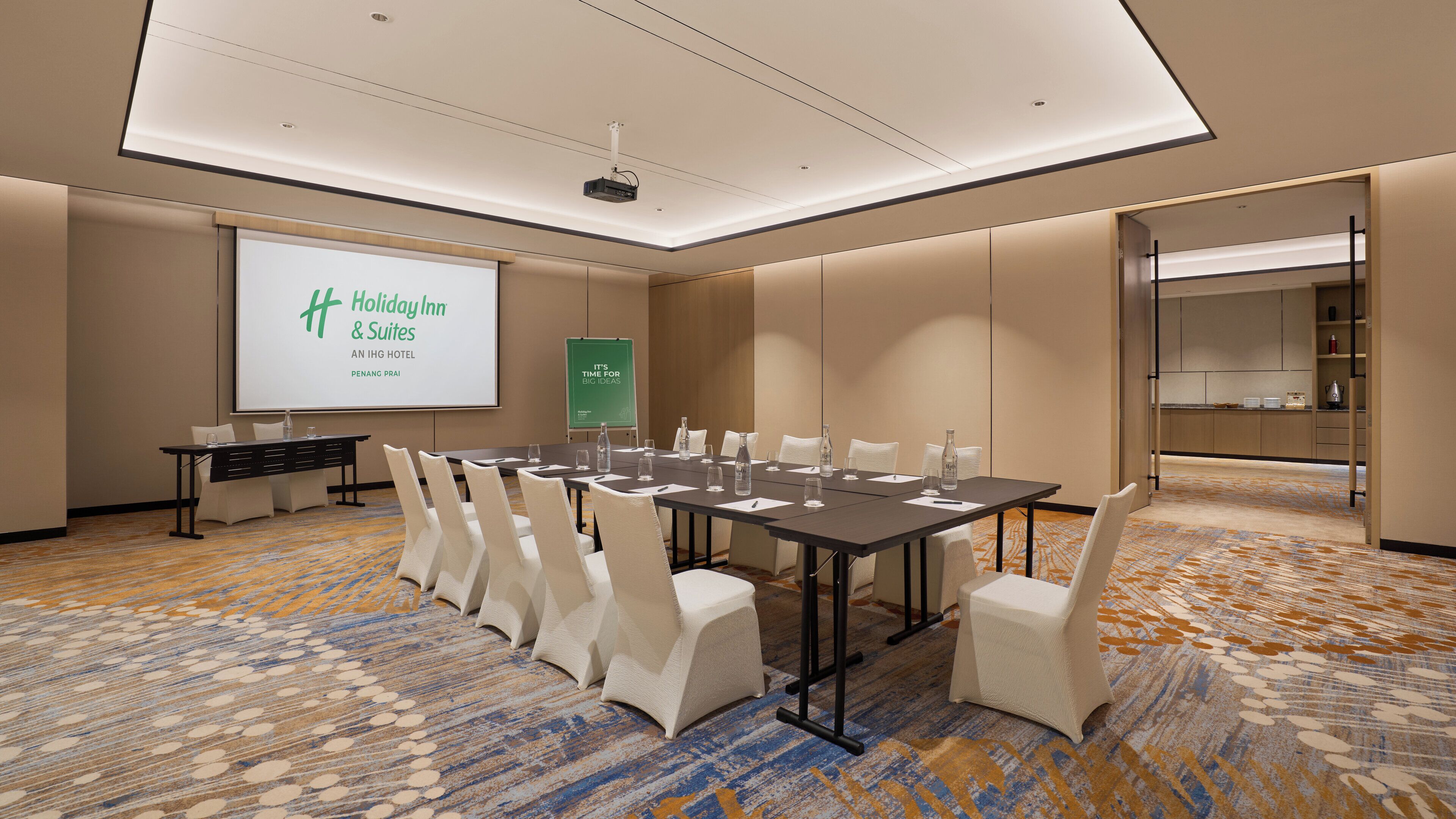 Un salón de conferencias con una larga mesa, sillas, una pantalla de proyector y una presentación sobre Holiday Inn & Suites.