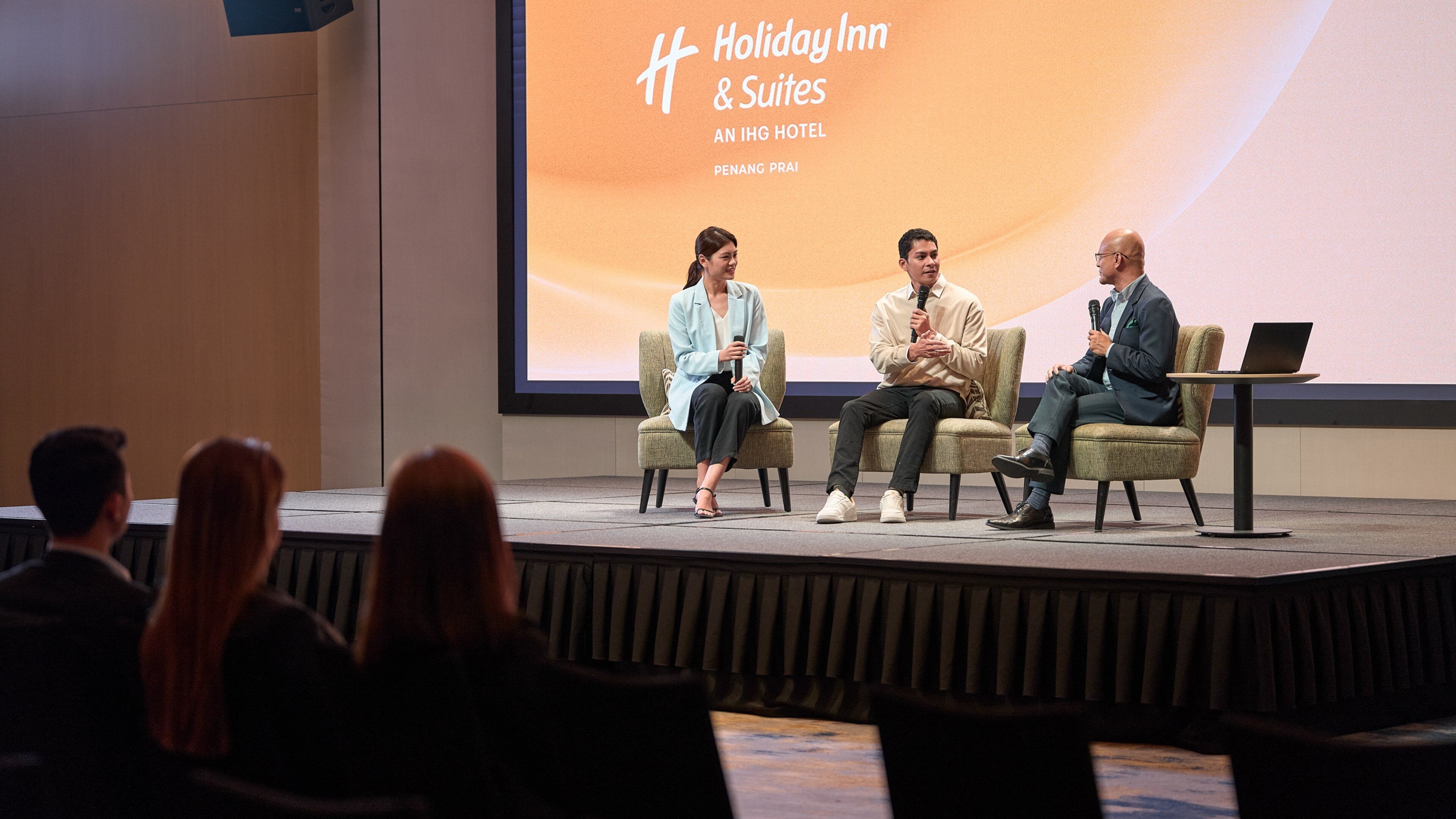 Panel de discusión con tres personas sentadas en un escenario, mirando a una audiencia. Una pantalla grande al fondo muestra el texto 'Holiday Inn & Suites AN IHG HOTEL PENANG PRAI'.