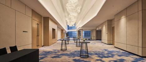 A hotel corridor with a modern design, featuring a long reception desk, multiple seating areas with tables, and a large, open space with a high ceiling and natural light from large windows.