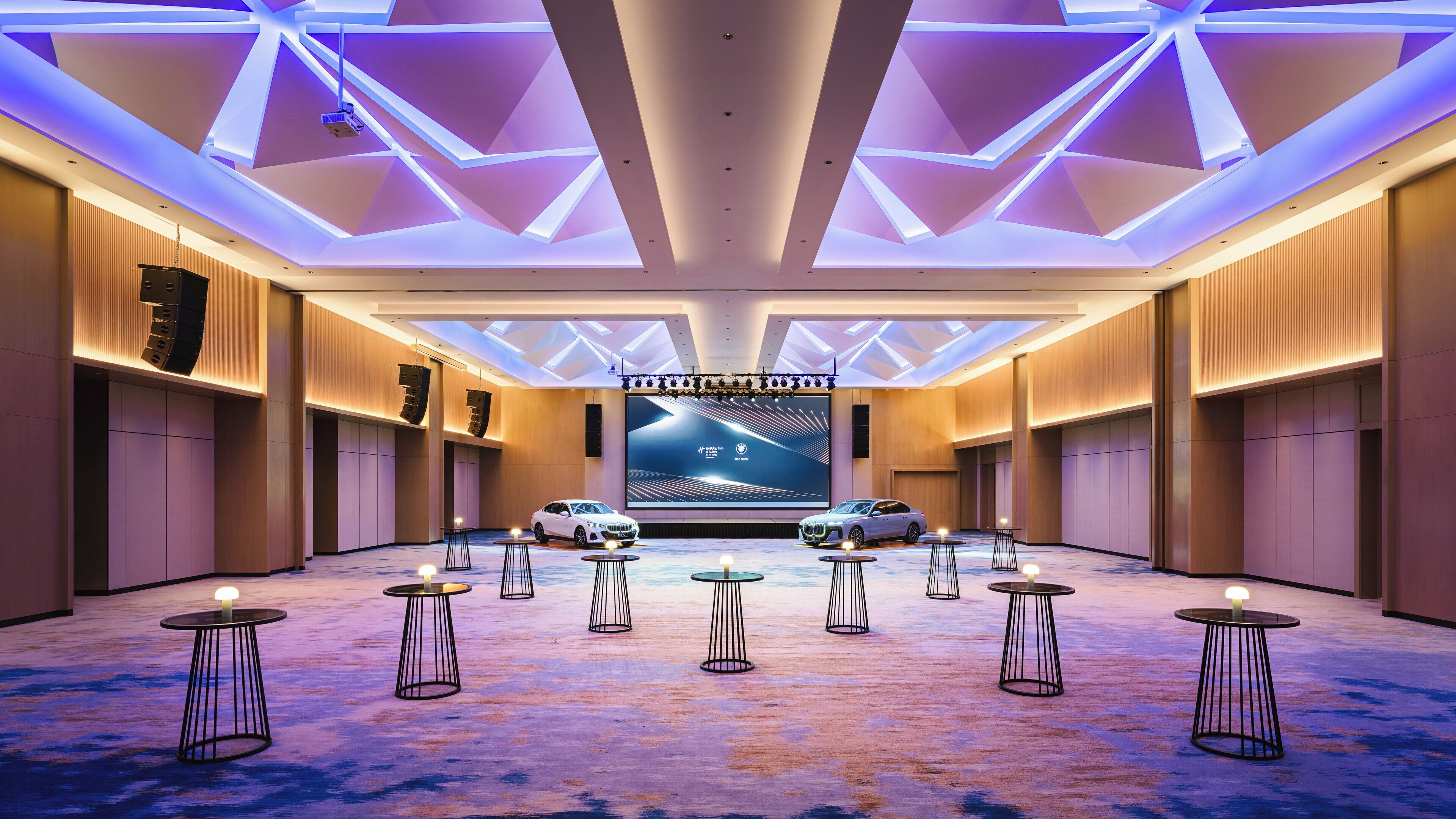 A spacious hall with a large screen displaying a car advertisement, multiple tables with candles, and a modern ceiling design.