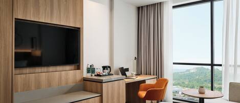 A modern hotel room with a large TV, a desk with a laptop, a chair, and a view of a mountainous landscape through large windows.