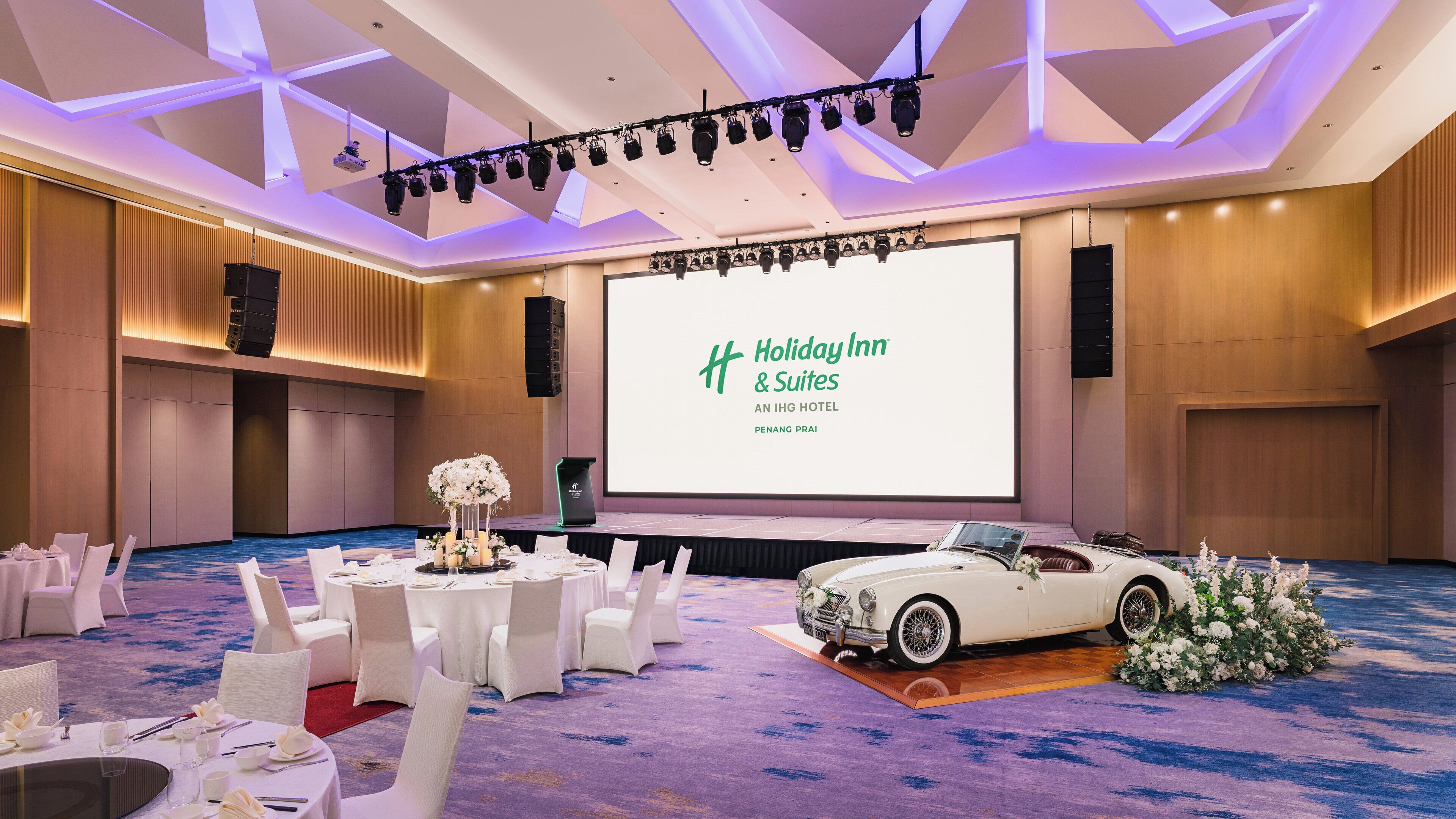 A conference room with a large screen displaying "Holiday Inn & Suites" branding, a white convertible car, and a floral arrangement on the carpet.