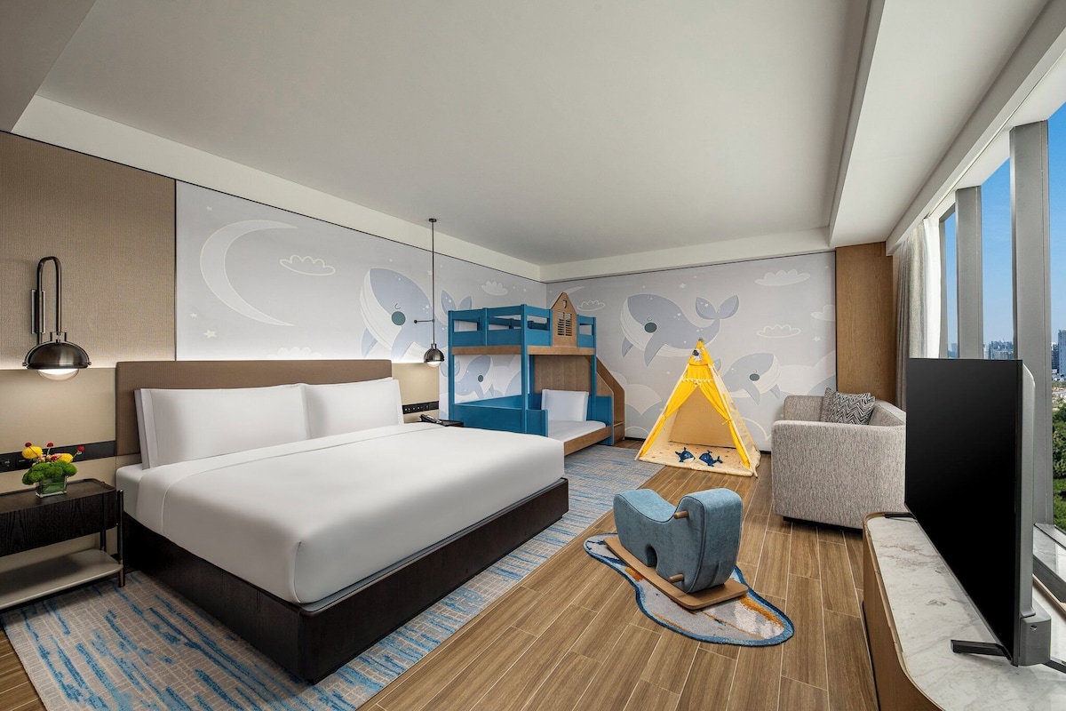 Una habitación de hotel moderna con una cama grande, un área de descanso con sofá y sillón, televisión y vista a la ciudad.