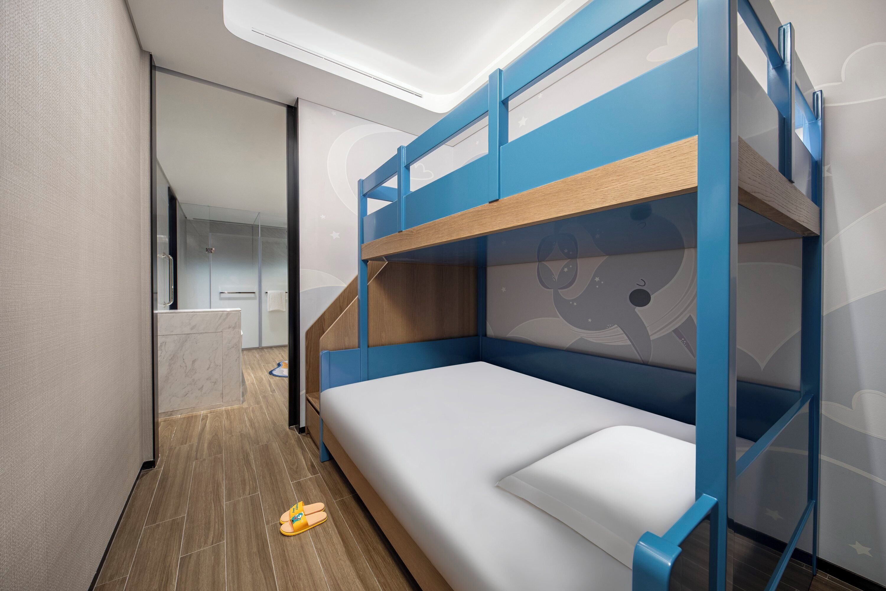 A bunk bed with a white mattress and blue frame, a wooden floor, and a bathroom with a white sink and mirror.