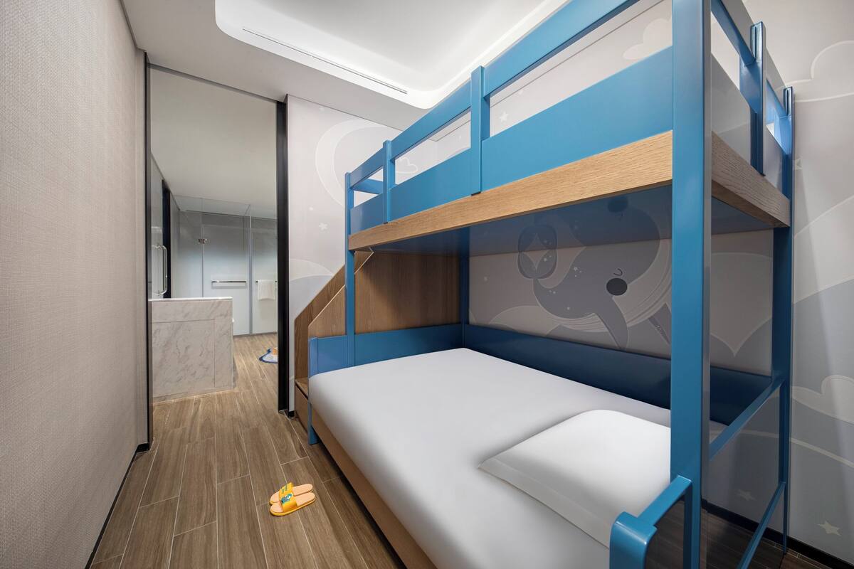 Una cama litera con colchón blanco y estructura azul, suelo de madera, y un baño con lavabo y espejo blancos.