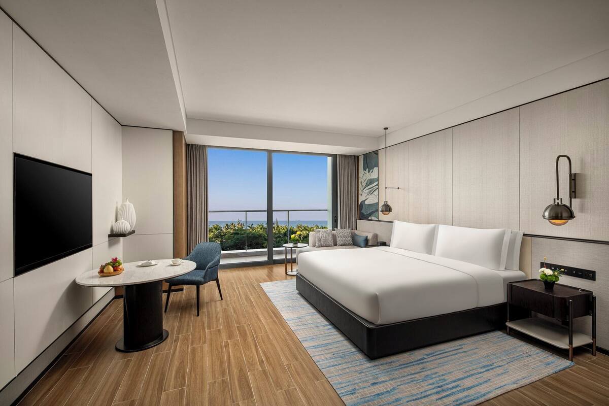 Una habitación de hotel moderna con una cama grande, televisión, una mesa de comedor y un balcón con vistas.