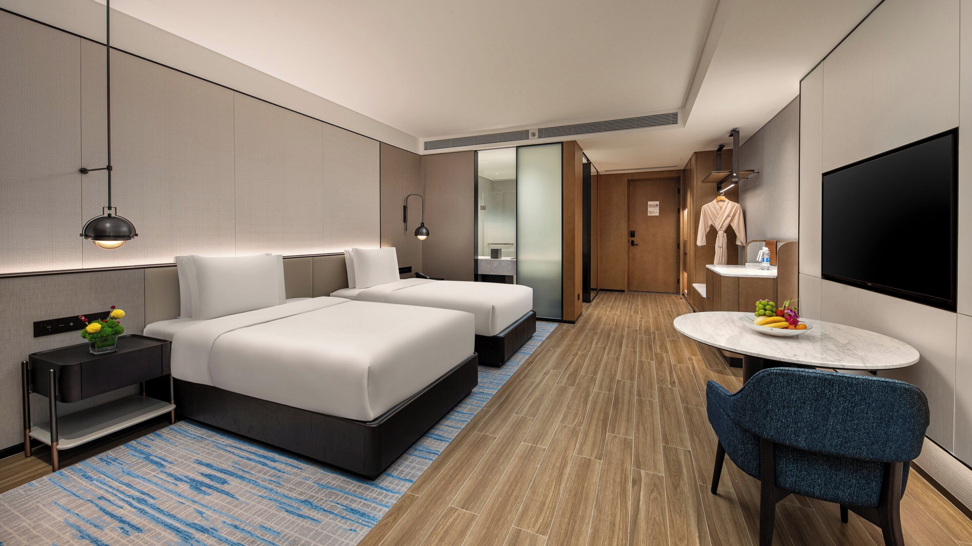 Ein modernes Hotelzimmer mit zwei Betten, einem Schreibtisch, einem Stuhl, einem Fernseher und einem kleinen Tisch mit Obst.
