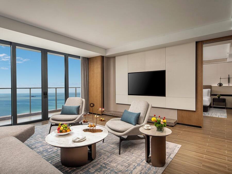 A modern hotel room with a large balcony, a flat-screen TV, and a dining area with a table set for two.