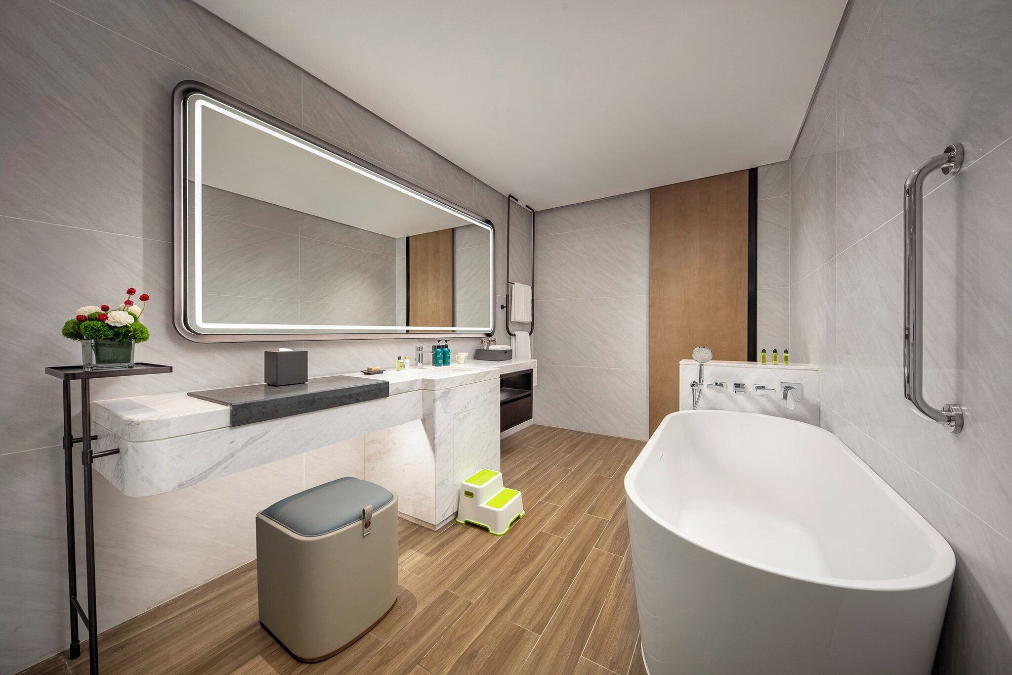 A modern bathroom with a freestanding bathtub, a large mirror, a vanity with a marble countertop, and a wooden floor.