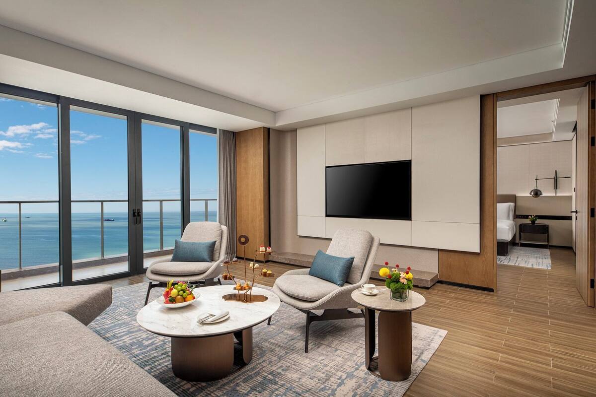 Una habitación de hotel moderna con un balcón grande, un televisor de pantalla plana y una zona de comedor con una mesa dispuesta para dos.