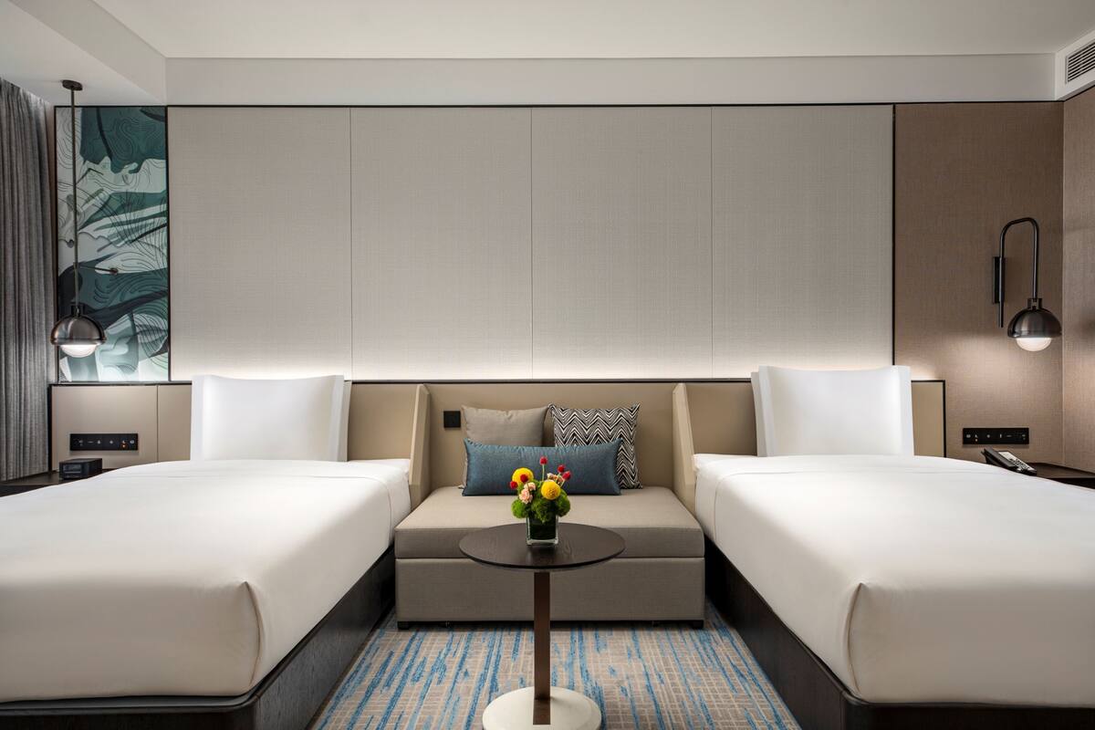 Habitación de hotel con dos camas, un sofá cama, una mesita con un jarrón de flores y un ventanal con cortinas.