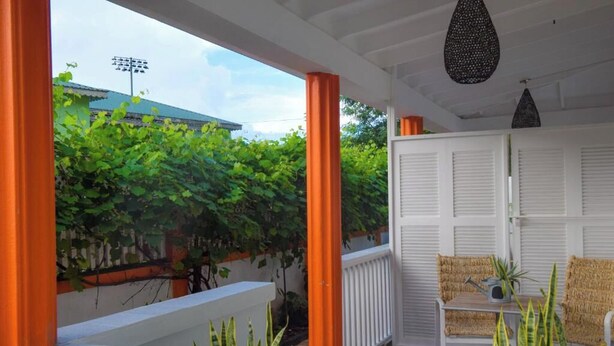 A covered outdoor seating area with a white door, orange columns, and a view of greenery and a building in the background. - Cozy studio in gorgeous Soufriere with WiFi, AC (Soufriere)