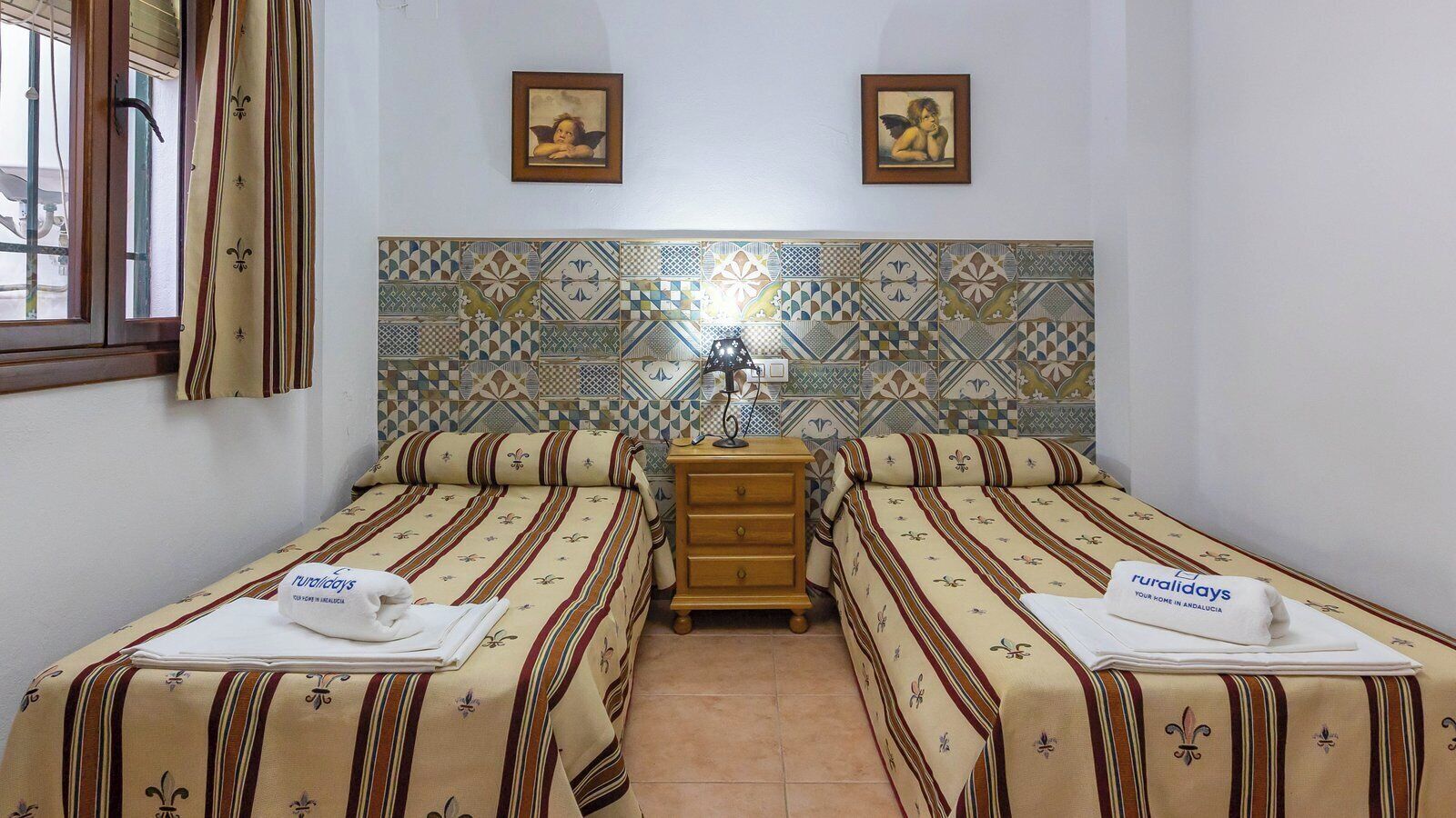 Habitació d'hotel amb dos llits, una tauleta de nit de fusta i una paret amb rajoles i un dibuix geomètric.