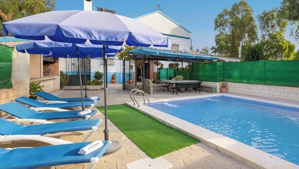 En swimmingpool med blå solstolar och parasoller, ett avskärmat sittområde och en byggnad i bakgrunden.