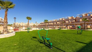 Un espace extérieur bien aménagé, doté d’équipements de musculation, de palmiers et, au fond, d’une rangée d’immeubles d’appartements modernes.