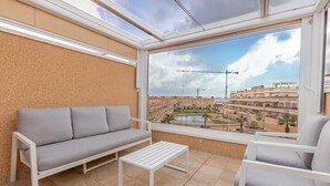 Un balcon avec un canapé gris, une table blanche et une vue sur un quartier résidentiel avec des immeubles et une piscine.