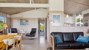 Een moderne woonkamer met een zwarte leren bank, een eettafel met stoelen, en een keuken met een wit aanrecht en hangende lampen.