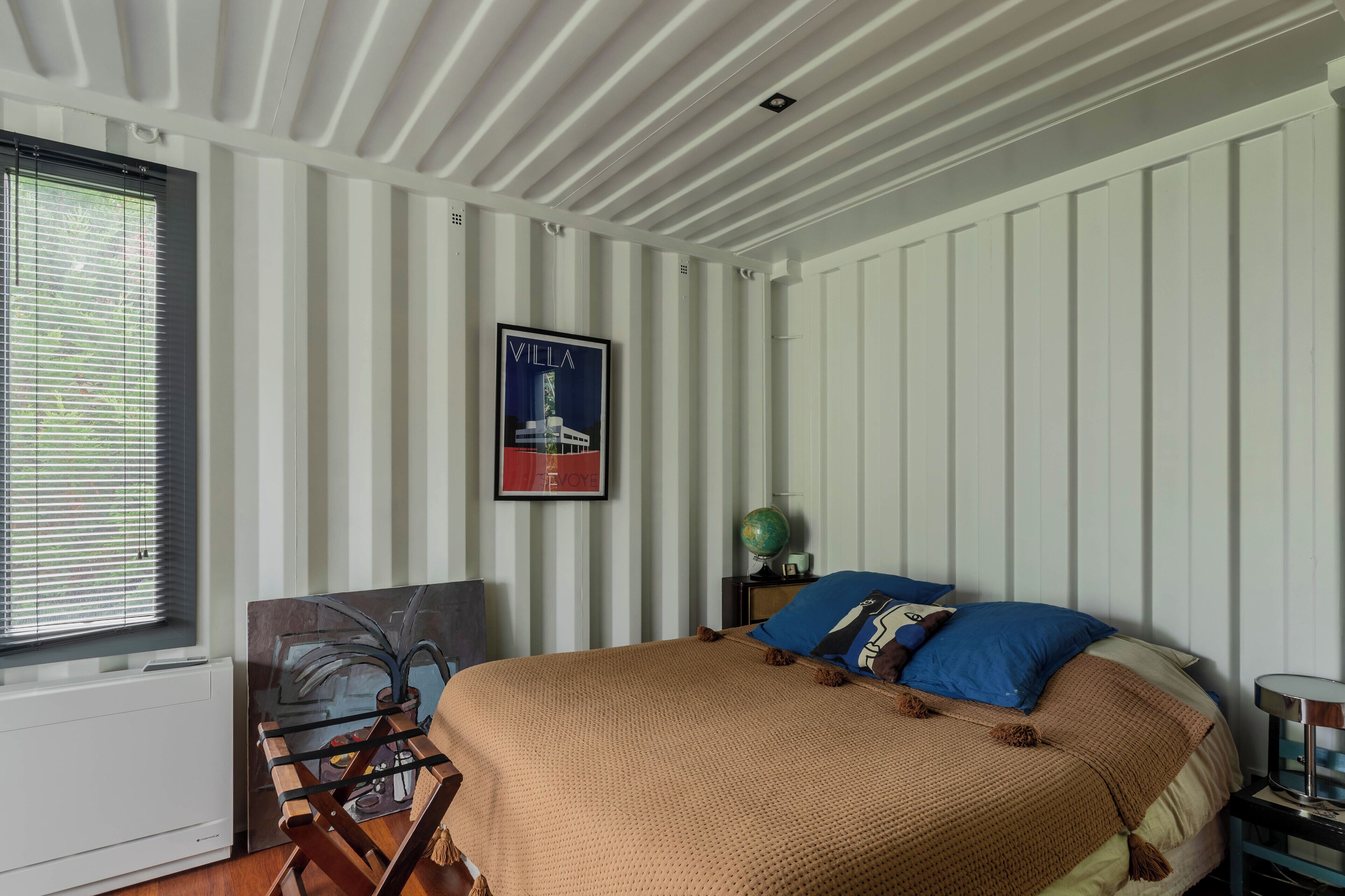 Une chambre à coucher avec un lit, un globe terrestre posé sur une table de chevet, un tableau encadré accroché au mur et une fenêtre avec des stores.