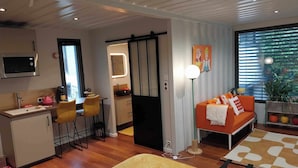 Una habitación de hotel moderna con kitchenette, zona de comedor, sofá y un amplio ventanal con persianas.