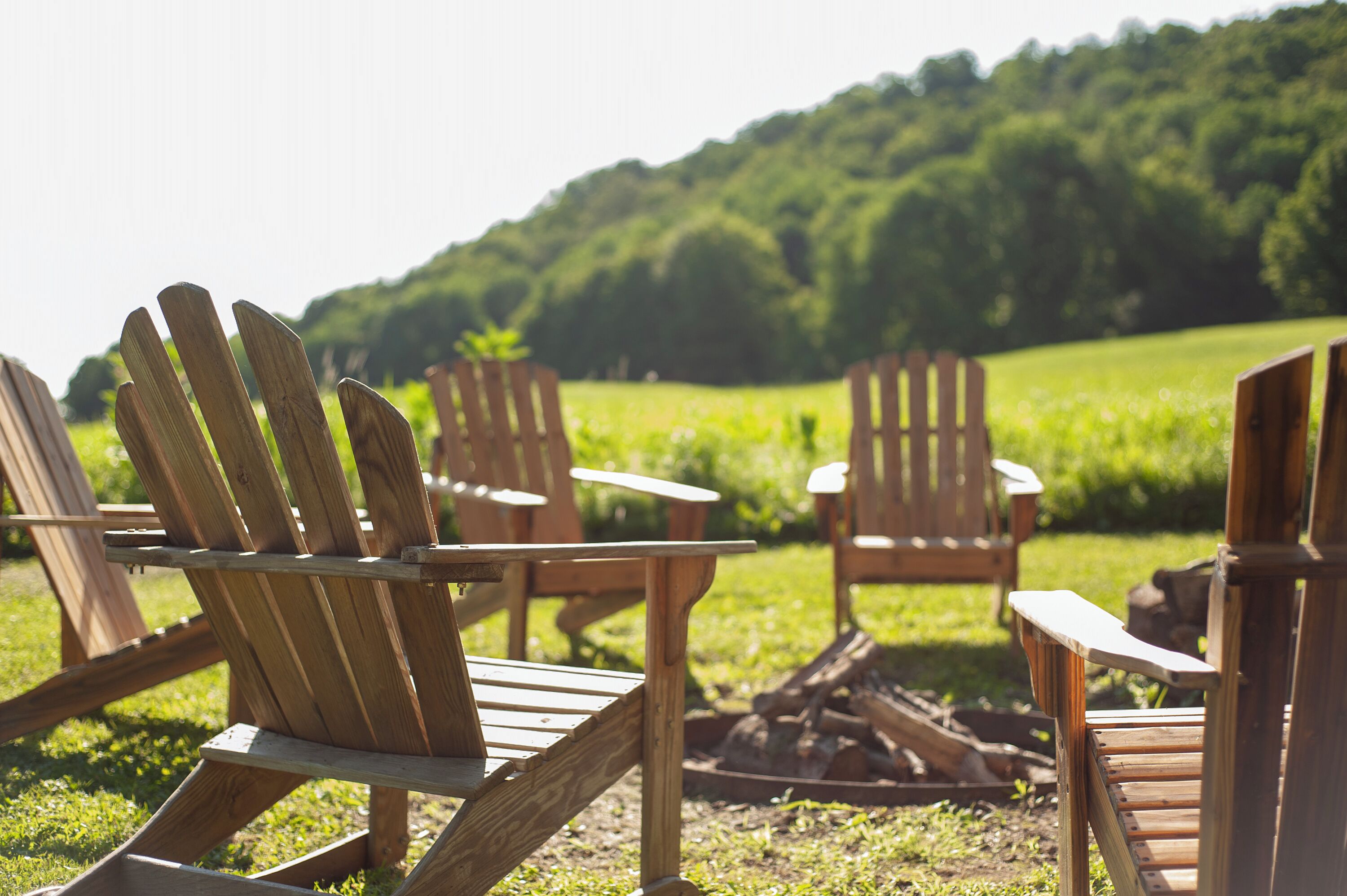 Outdoor wooden chairs and tables arranged on a grassy field with a forested hill in the background.