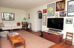 A living room with a sofa, a television, a bookshelf, and various framed artworks on the wall. - 4BR Catskills Farmhouse on 26 Private Acres. Mountain Views, Sleeps 8 (Jefferson)
