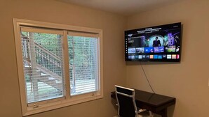 Quarto de hotel com uma janela grande, uma escrivaninha com cadeira e uma televisão de tela plana fixada na parede, exibindo diversos canais.