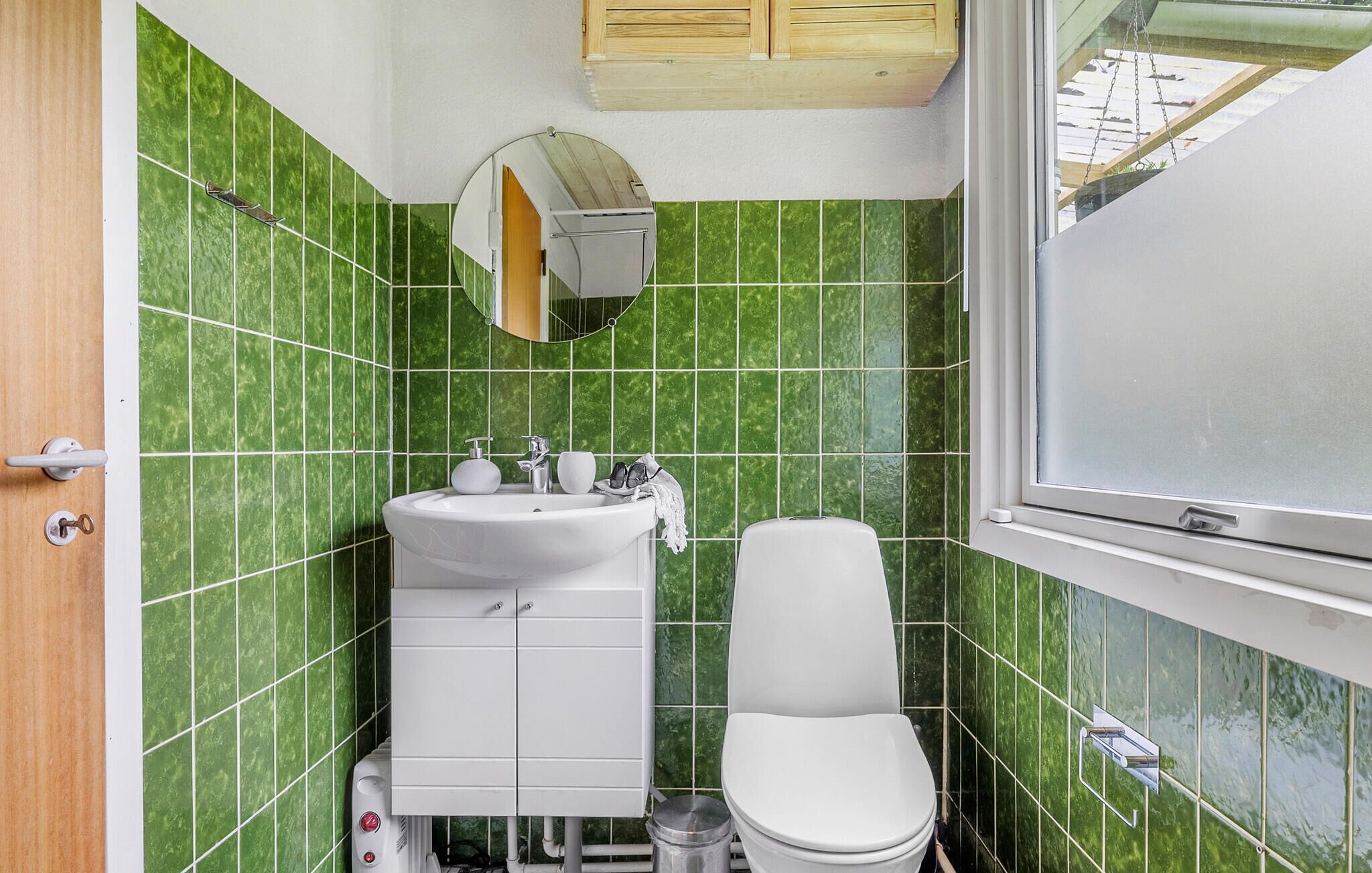 A small bathroom with green tiles, a white sink, a toilet, and a window.