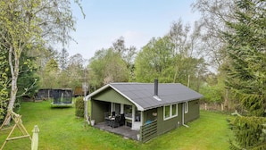 En liten, envåningsstuga med en veranda och en skorsten, omgiven av en gräsklädd trädgård och träd.