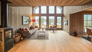 A spacious living room with a large fireplace, wooden flooring, and large windows offering a view of a lake. - Stunning home in Vinje with WiFi (Vinje)