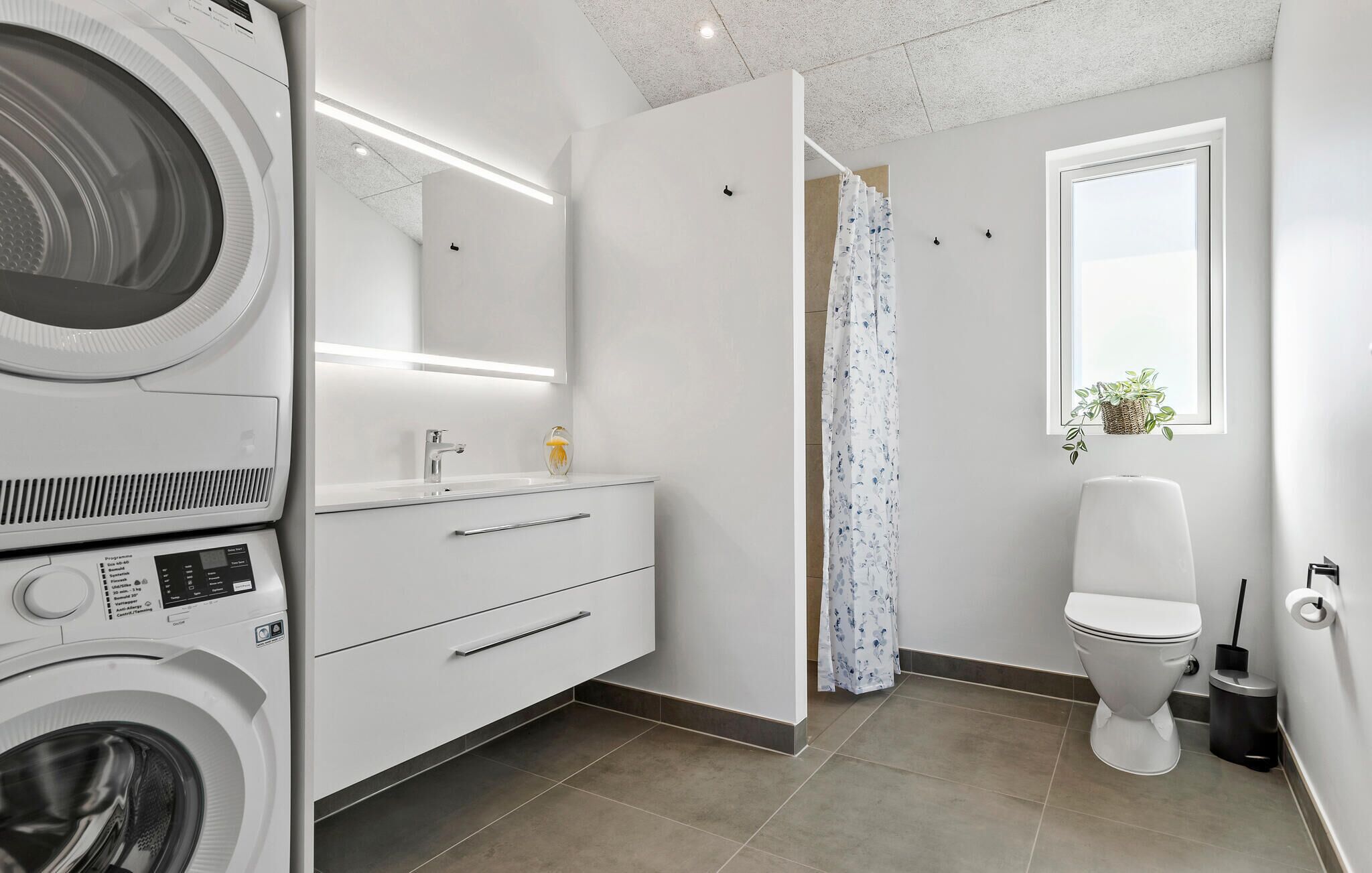 Ein kleines, modernes Badezimmer mit integrierter Waschmaschine, einem Waschtisch mit Spiegel, Toilette und einem Fenster mit Pflanze.