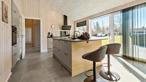 A modern kitchen with a central island, a dining area with two bar stools, and a large sliding glass door leading to a view of a parked car and trees.