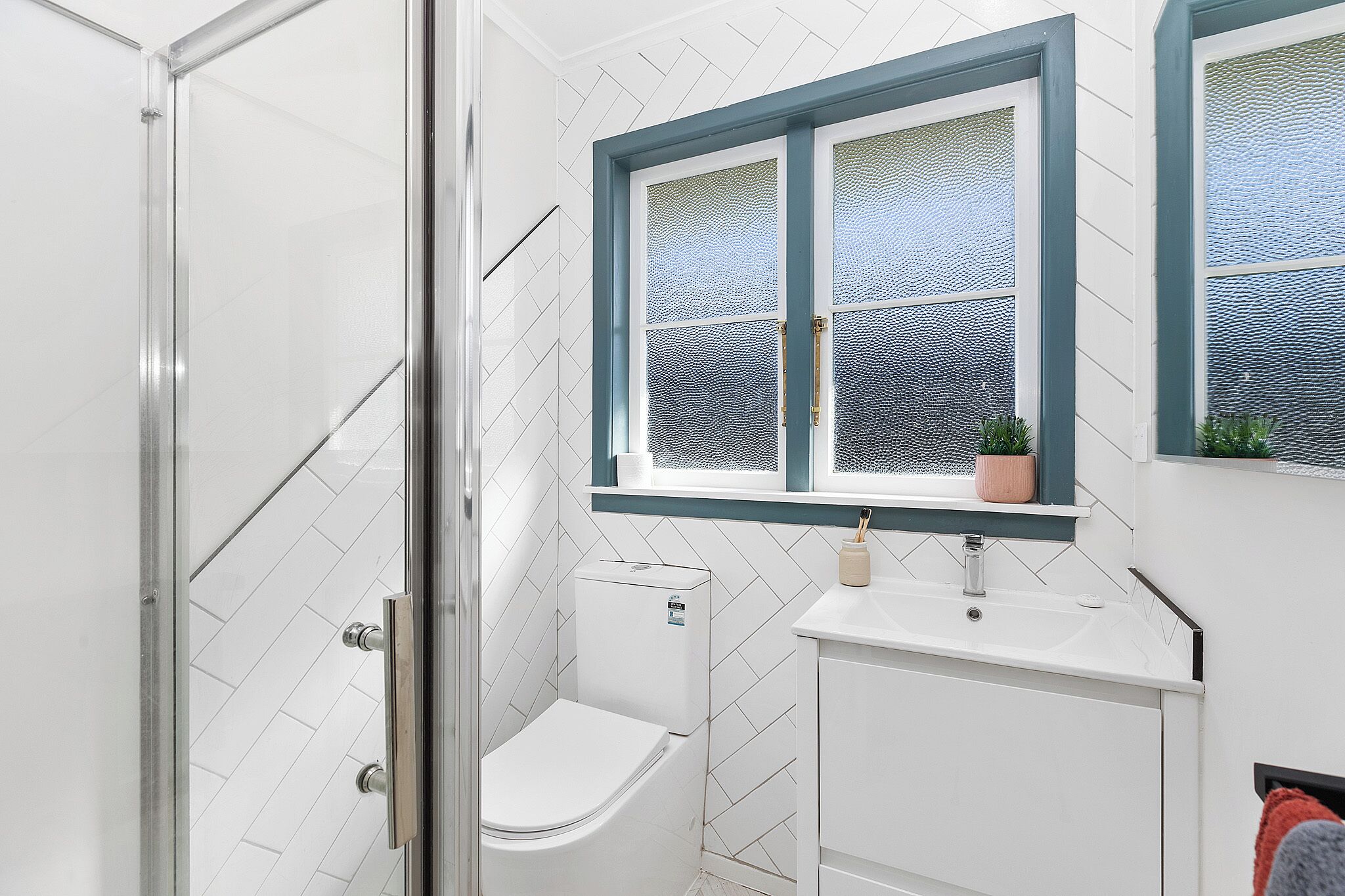 A bathroom with a glass shower enclosure, a toilet, a sink, and a window with a blue frame.