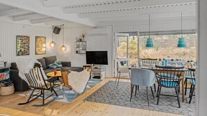 Een moderne woonkamer met een bank, een eethoek met stoelen en een tafel, een televisie en een groot raam met uitzicht op de omgeving.