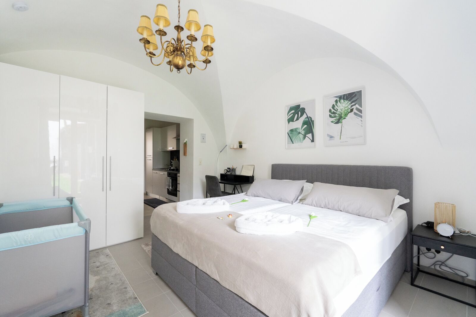 A modern bedroom with a large bed, a desk, and a kitchenette in the background.