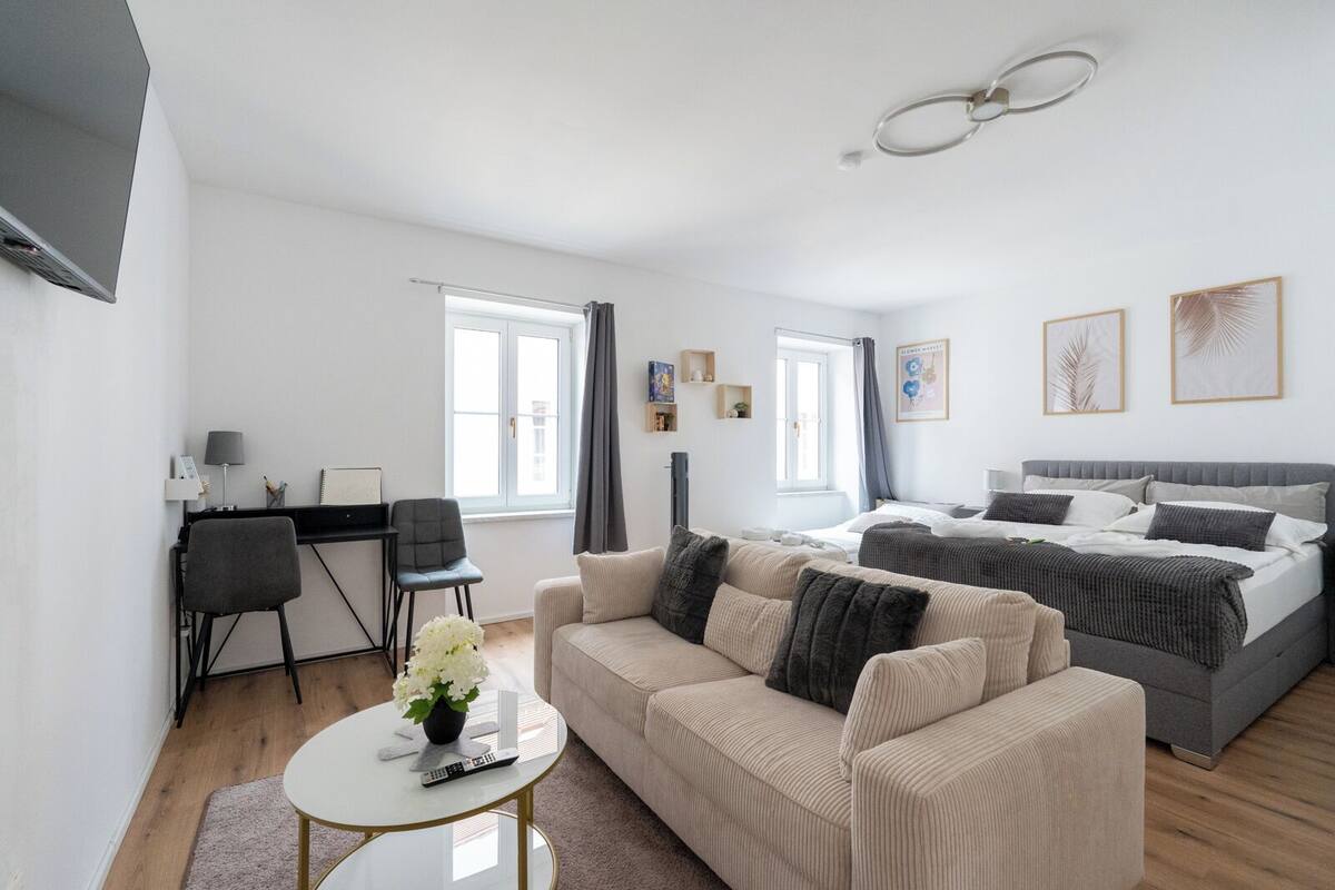 Ein modernes, helles Schlafzimmer mit einem großen Bett, einem Schreibtisch, einem Sofa und einem kleinen Tisch mit einer Fernbedienung.