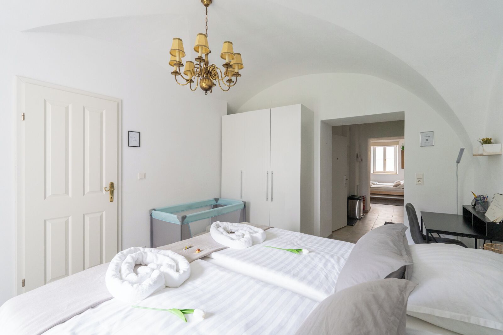 Een hotelkamer met twee bedden, een bureau, een stoel, een kroonluchter en uitzicht op de gang.