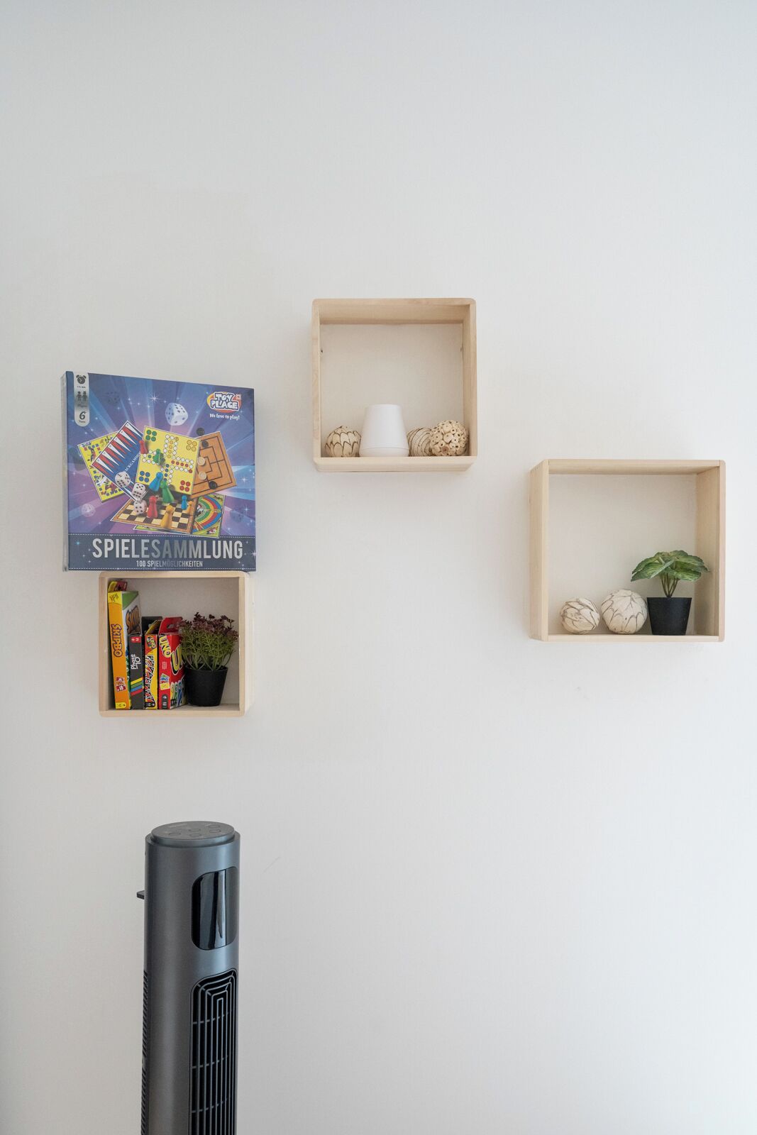 Cuatro estantes de madera en una pared, cada uno con objetos diferentes: un juego de mesa, una taza, una planta en maceta y adornos.