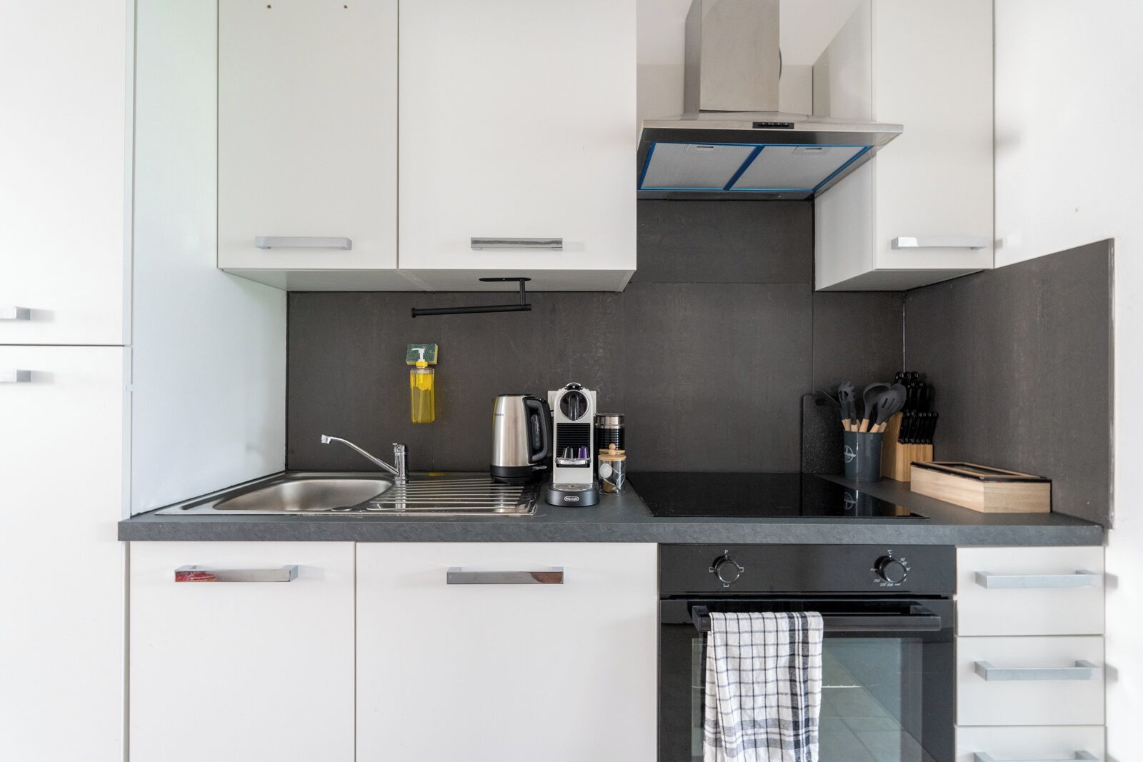 A modern kitchen with white cabinets, a black countertop, a stainless steel sink, a coffee machine, and a range hood.